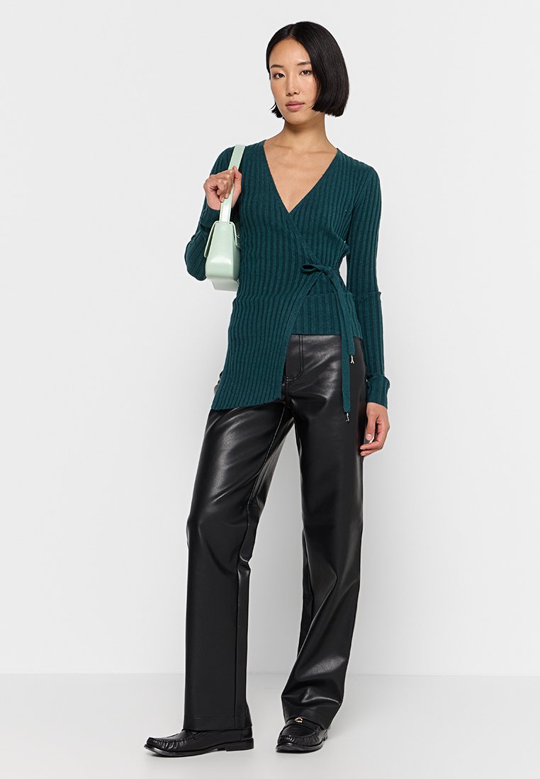 Top avvolgente a coste color teal con maniche lunghe, abbinato a pantaloni in pelle nera a vita alta e una piccola borsa verde menta. Presenti dettagli testurizzati.