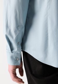 Camisa de manga larga en azul claro, hecha de una tela suave, con puños abotonados y un dobladillo recto. Pantalones negros visibles en la parte inferior.