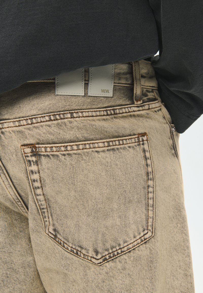 Nahaufnahme der hinteren Tasche und des Bunds einer beige-farbenen Jeans aus Denim mit einem weißen Leder-Brandpatch, getragen mit einem schwarzen Langarmshirt.