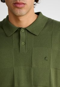 Olivengrønn polo-skjorte med et strukturert rutete mønster, som har krage og to knapper, samt en liten preget logo på brystet.