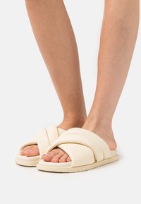 Ljust beige slide sandaler med en flätad, vadderad design. Mjukt material och tjock sula, som visar tårna, mot en enfärgad bakgrund.