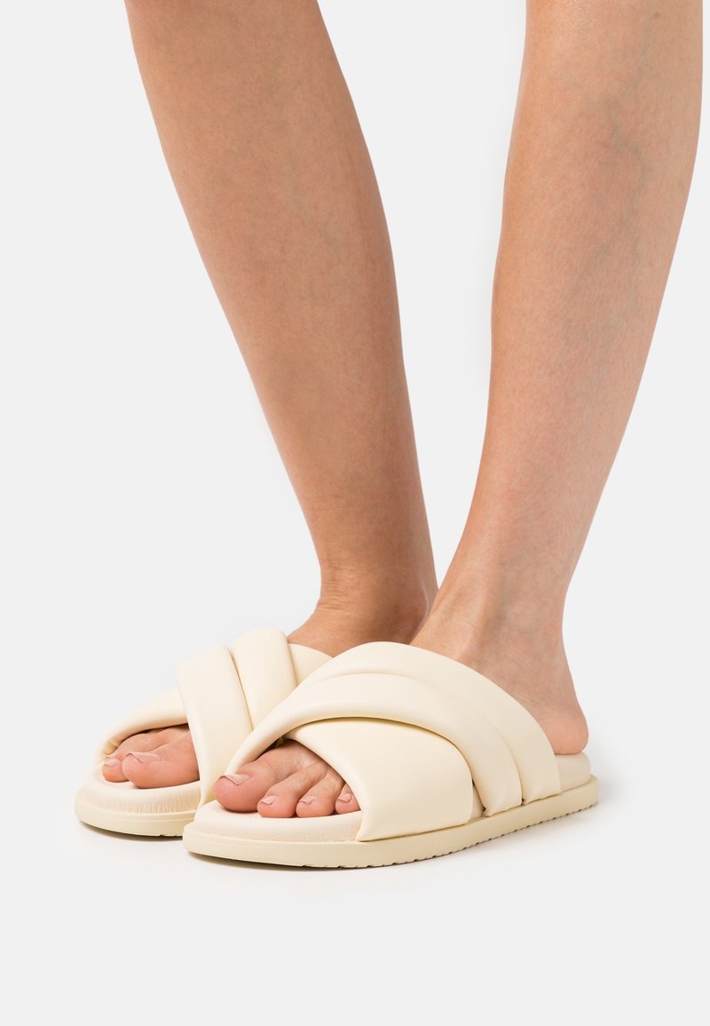 Ljust beige slide sandaler med en flätad, vadderad design. Mjukt material och tjock sula, som visar tårna, mot en enfärgad bakgrund.