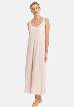 Vrouw met blond haar die een mouwloze, lichtroze maxi-jurk draagt met een klein borstzakje, blootsvoets staand tegen een effen achtergrond.