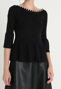 Czarny top peplum wykonany z materiału dzianinowego, z rękawami o długości trzech ćwierci i perłowym akcentem przy dekolcie, zestawiony z gładką, czarną skórzaną spódnicą.