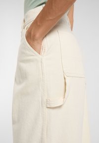 Campione di tessuto color crema con superficie texture, caratterizzato da una tasca laterale e un design dalla vestibilità casual, mostrato da un angolo.