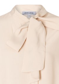 Beige Bluse aus glattem Stoff mit Stehkragen und einer großen, gebundenen Schleifendetail am Hals. Etikett zeigt die Marke "MORE & MORE."