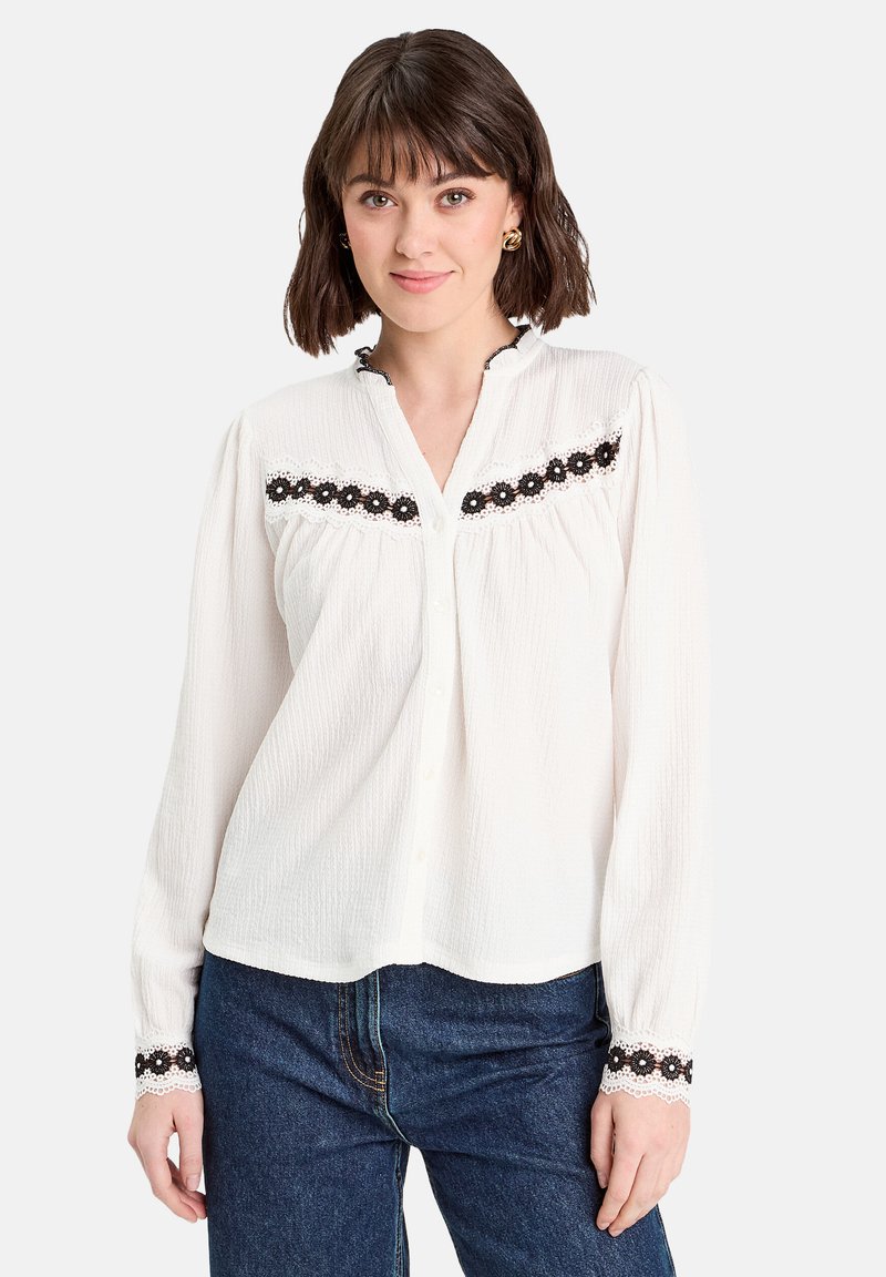 Blouse blanche à manches longues en tissu texturé, boutonnée à l'avant, avec des accents en dentelle florale noire sur la poitrine et les poignets, et un col festonné.