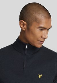 Navy zip-neck sweater made of knitted fabric, featuring a ribbed collar and a small gold logo embroidered on the left chest.