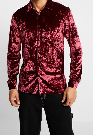 Mand iført en langærmet, knappet, knust fløjls skjorte i dyb rød med sorte bukser mod en ensfarvet baggrund.