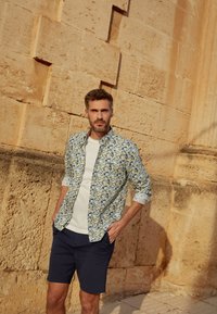Chemise à boutons avec un motif floral en jaune, bleu et vert, portée sur un t-shirt blanc, associée avec un short marine. Fond en pierre texturée.