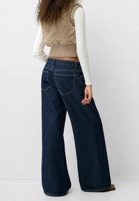 Hellebeiner Jeans aus dunklem Denim mit weitem Bein und hoher Taille, ausgestattet mit zwei Gesäßtaschen und kontrastierenden Nähten. Kombiniert mit einem beigem, strukturierten Weste.