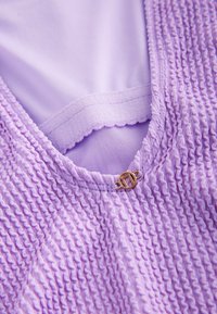 Lavendrowa tkanina o fakturze z falbankami i gładką wewnętrzną podszewką. Przy dekolcie znajduje się małe zapięcie w odcieniu złota.