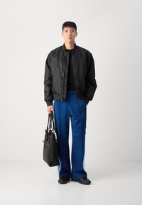Chaqueta bomber negra con cremallera, camiseta negra, pantalones anchos azules y zapatillas negras. Lleva un bolso negro con un diseño acentuado.