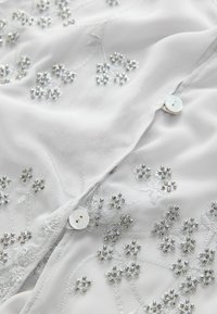 Tissu blanc avec broderie florale délicate en perles et fils argentés, avec des boutons nacrés sur un vêtement plié.