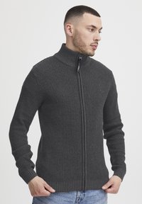 Pulover cu fermoar de culoare gri cărbune, cu un model texturat, guler înalt și mansete și tiv ribbate, purtat cu blugi de denim albastri.