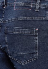 Vaqueros de mezclilla en azul oscuro con una ligera textura. Presentan un bolsillo trasero con costura rosa y un parche rectangular encima de él.