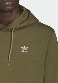 Olivově zelená mikina s kapucí z měkkého materiálu a přední kapsou ve tvaru klokaního vaku. Na hrudi je bílý logo Adidas a kontrastní šňůrky na stažení.