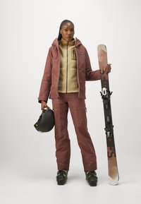 Skijakke i rødbrun, fleece-foret indvendigt; beige fleece halv-zip under. Sort skihjelm, ski med bindinger.