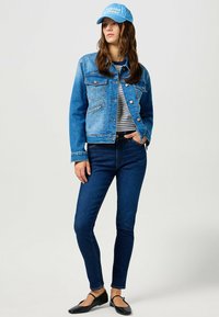 Denim jacket vaaleassa ja tummassa sinisessä, taskut mukana, yhdistettynä raidalliseen paitaan ja tummiin farkkuihin. Lisävarusteet sisältävät sinisen lippiksen ja mustat ballerinat.