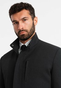 Cappotto nero con texture, colletto alto, chiusura con zip frontale e tasca discreta. Indossato sopra una camicia con colletto e cravatta.