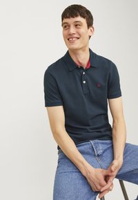 Mörkblå polotröja med röd krage, treknapplåsning och en broderad logotyp på bröstet. Bärs tillsammans med ljusblå denimjeans.