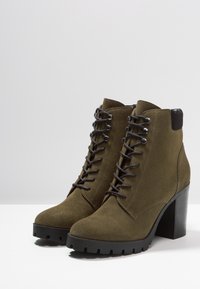 Olivengroene suède enkellaarzen met een hoge blokhak, veterfront en contrasterende zwarte accenten bij de hak en de vetergaatjes.