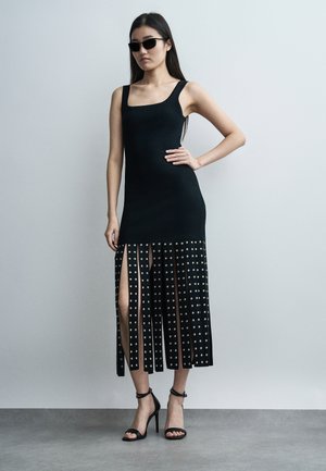 Kobieta w czarnej sukience bez rękawów z rozcięciem i srebrnymi ćwiekami, w czarnych sandałach na wysokim obcasie i czarnych okularach przeciwsłonecznych, stoi na tle gładkiej ściany.