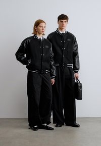 Jachete bomber negre cu mâneci din piele, manșete albe zimțate, asortate cu pantaloni negri largi, un model poartă o geantă neagră. Fundal neutru.