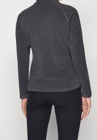 Persona vestida con una camiseta de manga larga ajustada de color gris oscuro y leggings negros, mostrada de espaldas frente a un fondo sencillo.