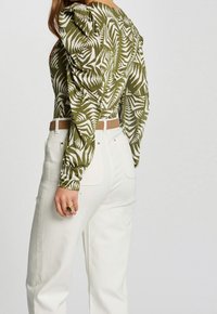 Haut à manches longues à motifs vert olive avec épaules bouffantes, associé à un pantalon blanc taille haute et une ceinture beige. Bague en or à la main.