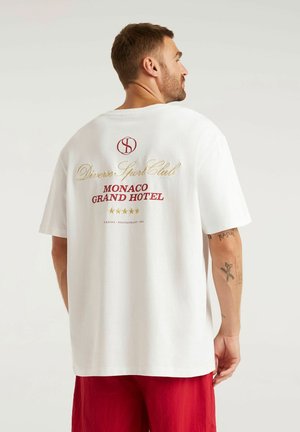 Mężczyzna ubrany w białą koszulkę z napisem „Diverse Sport Club Monaco Grand Hotel” oraz czerwone spodenki, przedstawiony od tyłu na jednolitym tle.