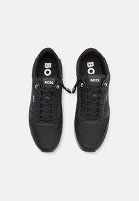 Zapatillas negras con parte superior sintética lisa, cierre de cordones y marca visible. Presentan un detalle de cremallera en el lateral y suela blanca.