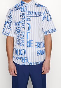 Kortärmad skjorta med krage i vitt med vertikala blå ränder. Har djärva blå textmönster och vita knappar.