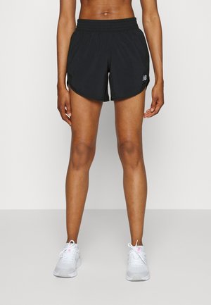 Atletisk person iført sorte løbeshorts og hvide sneakers, stående foran en ensfarvet lys baggrund.