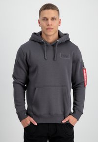 Sudadera con capucha gris que presenta un bolsillo delantero, cordones y un bolsillo con cremallera en la manga con una etiqueta roja. Textura suave, ajuste casual.