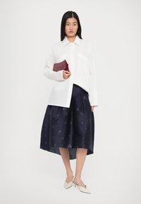 Woman wearing a white oversized shirt, navy patterned midi skirt, white heels, and holding a small maroon clutch purse.