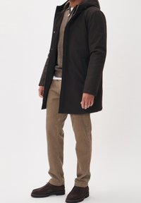 Man som bär svart huva jacka, bruna kordfloddsbyxor, beige tröja och mörkbruna mockaskor, står mot en enkel bakgrund.