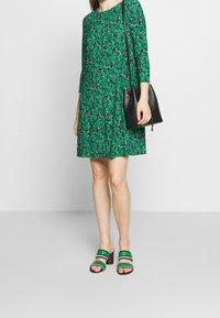 Green floral dress with three-quarter sleeves, a loose fit, paired with black bucket bag and multicolored block-heel sandals.