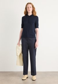 Polo de marină cu mânecă scurtă, cu guler cu nasturi, asortat cu pantaloni închiși la culoare și o jachetă bej deschis, purtat cu pantofi loafers bej pe un fundal neutru.