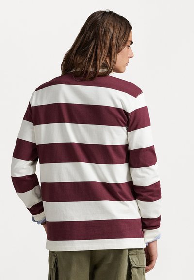 Persoană cu părul brunet de lungime medie, purtând un tricou de rugby cu mânecă lungă, cu dungi orizontale late maronii și albe.