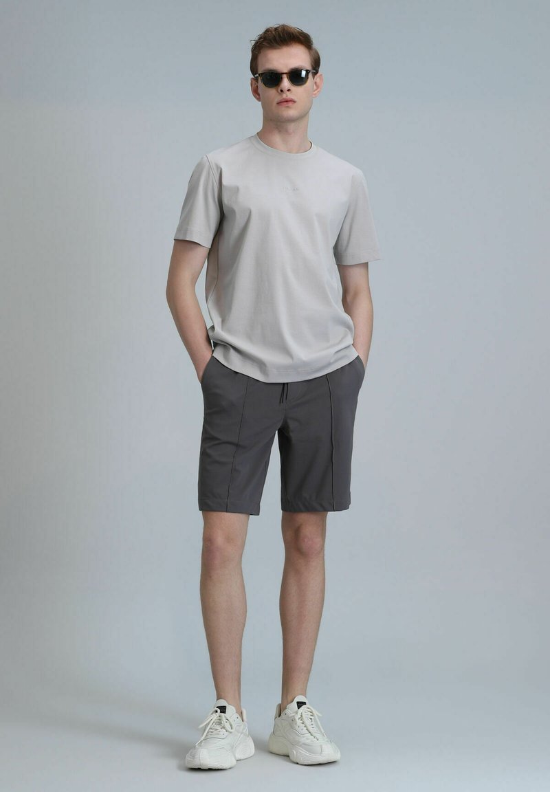 T-shirt gris clair à manches courtes avec un col rond et un logo subtil, associé à un short gris foncé et des baskets blanches, se tenant devant un fond uni.