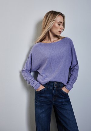 Lila Langarmoberteil mit lockerer Passform und aufgerollten Ärmeln, kombiniert mit dunkelblauen Jeans aus Denim mit doppeltem Knopfverschluss.