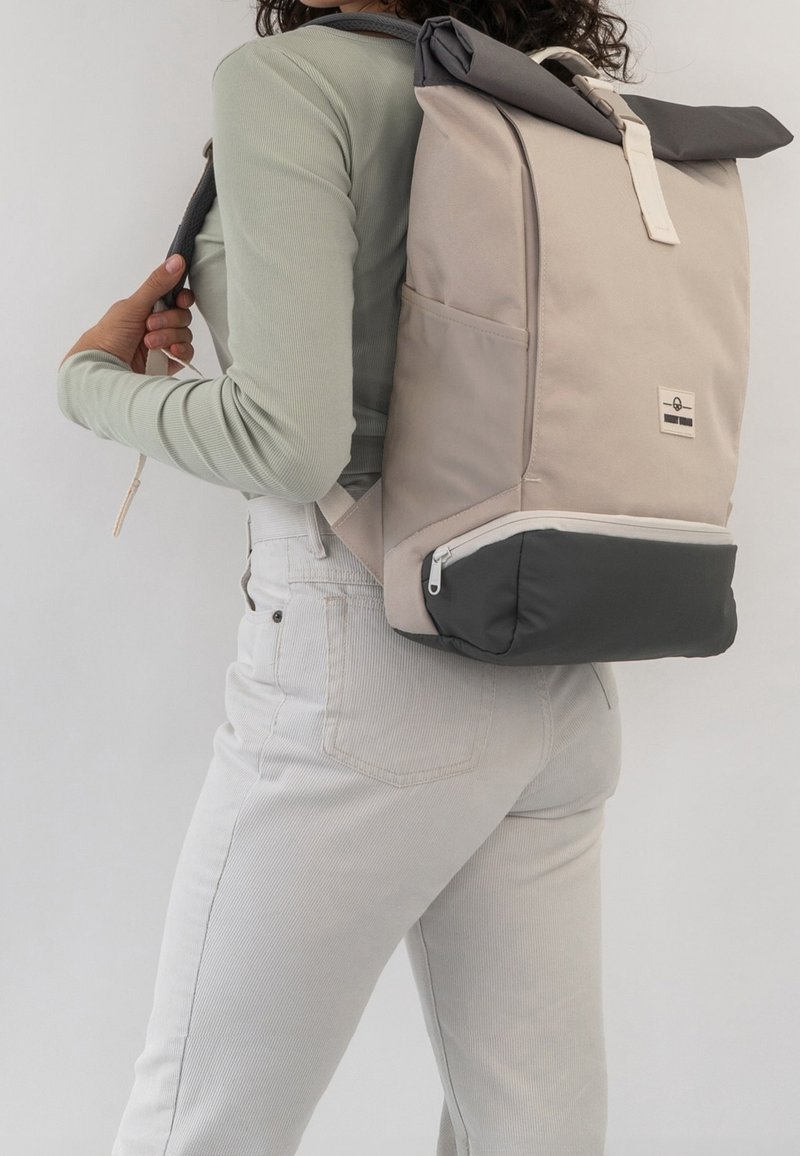 Personne portant un pantalon gris clair et une chemise verte à manches longues, portant un sac à dos beige et gris foncé à fermeture roulante sur l’épaule.
