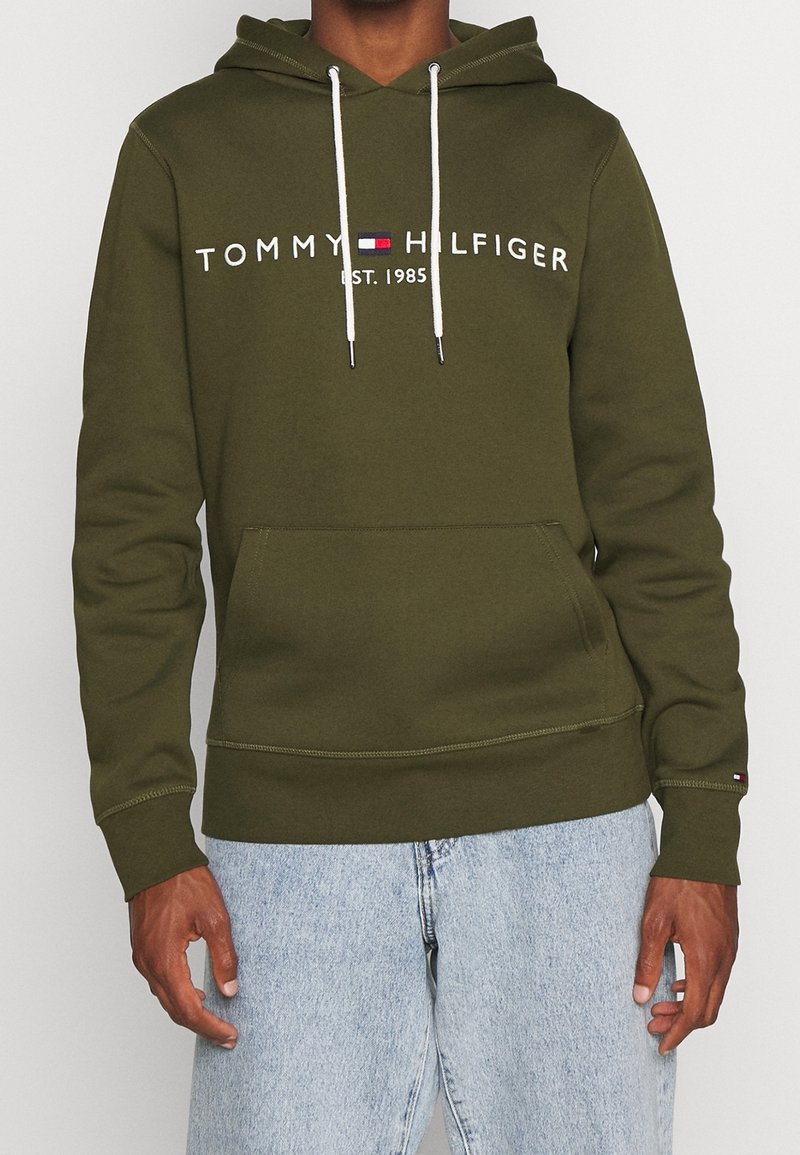 Hombre con sudadera oliva de Tommy Hilfiger con cordones blancos y jeans azul claros, de pie contra un fondo liso.