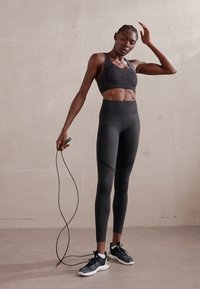 Sujeción deportiva negra y leggins de cintura alta con una textura suave y un diseño ajustado. La modelo sostiene una cuerda para saltar, vistiendo zapatillas negras.