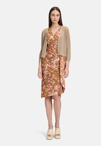 Braunblumiges Kleid mit rosafarbenen Akzenten, drapiertem Stil, kombiniert mit einem beigen Rippstrick-Cardigan. Das Model trägt cremefarbene Plateausandalen.