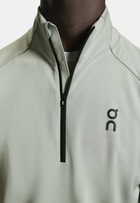 Hellgraues sportliches Viertel-Reißverschluss-Oberteil aus strukturiertem Stoff, mit schwarzem Reißverschluss und gedrucktem Logo auf der linken Brust.