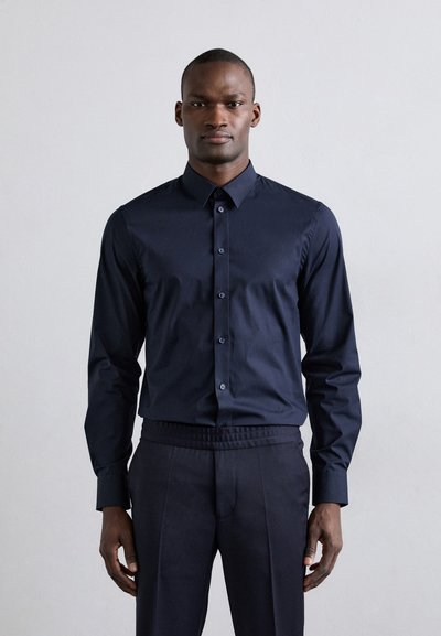 Camisa azul marino de botones, con mangas largas y cuello clásico, hecha de tela suave. Combinada con pantalones oscuros que tienen una cintura elástica.