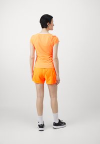 Top desportivo de cor laranja brilhante e calções, feitos de tecido que absorve a humidade. Apresenta mangas curtas, um design ajustado, e é acompanhado por sapatilhas de desporto pretas.