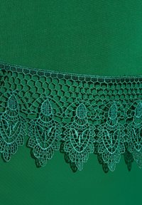 Smaragdgrüner Stoff mit aufwendiger Spitzenborte in gewelltem Design am unteren Rand, die detaillierte Muster und Strukturen zeigt.